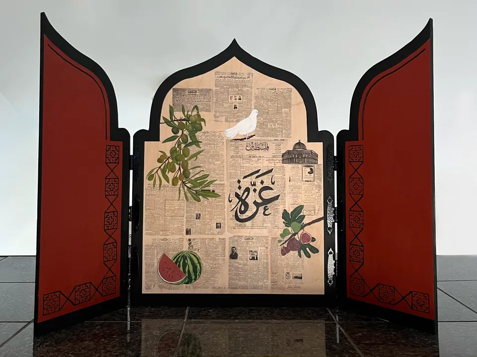 Structure en bois en forme de porte à trois volets : les battants rouges s’ouvrent sur des journaux palestiniens transférés, peints d’une colombe, de branches d’olivier, d’un dôme, d’une pastèque, de figues et de la calligraphie « Gaza ».